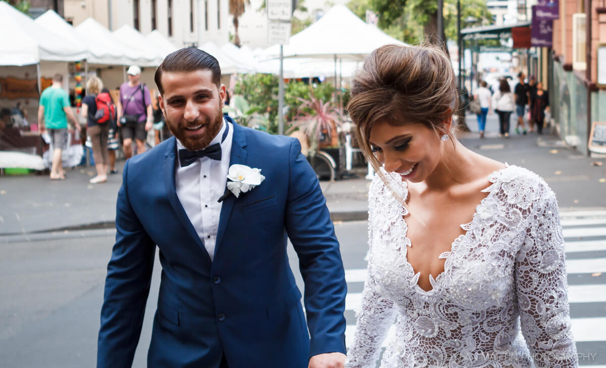 Sam and Ronda walk across the street in The Rocks, Sydney, Australia during their wedding day outside while there is a market/festival happening in the streets. | Photo Taken in Sydney, Australia by Dylan Martin Photography - London Ontario Wedding Photographer