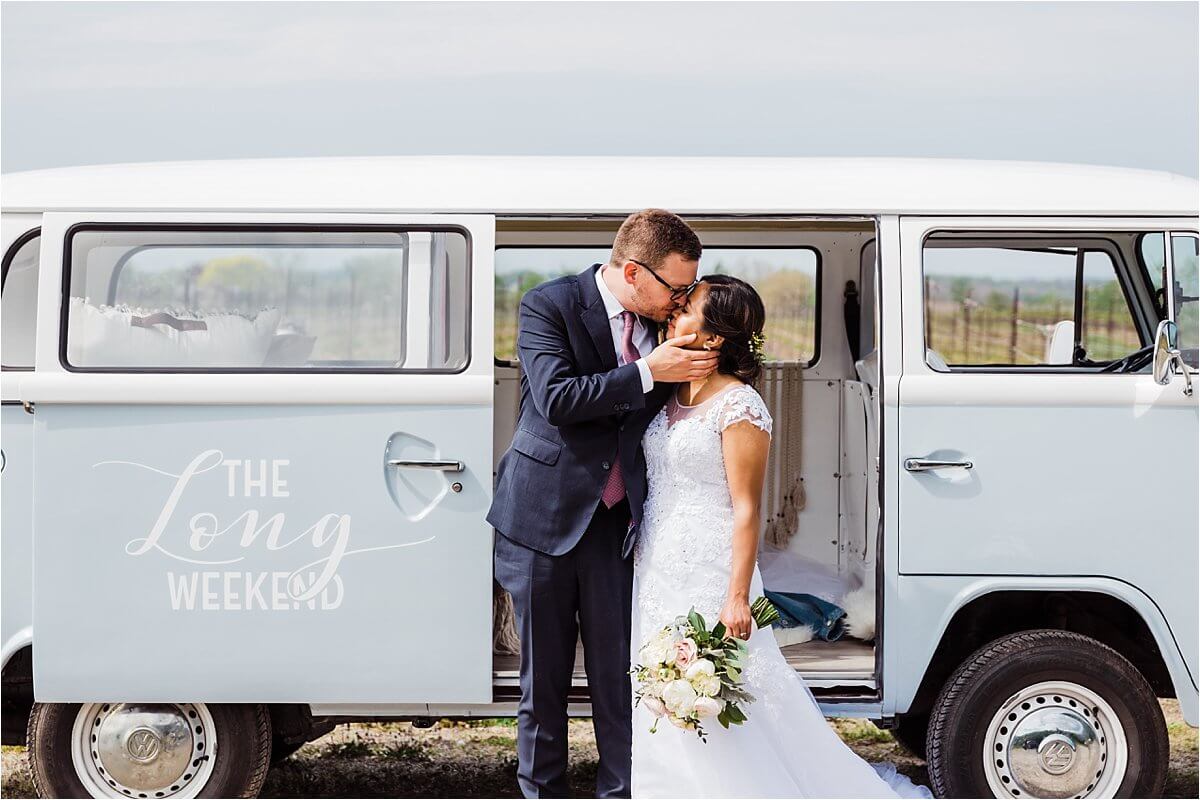 Groom kisses his bride softly in front of their wedding day car. A VW Kombi Van
