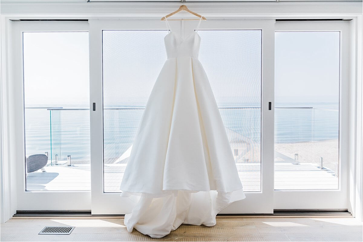 Bride's wedding dress hanging in the window overlooking the beach at their family cottage in Zurich, ON