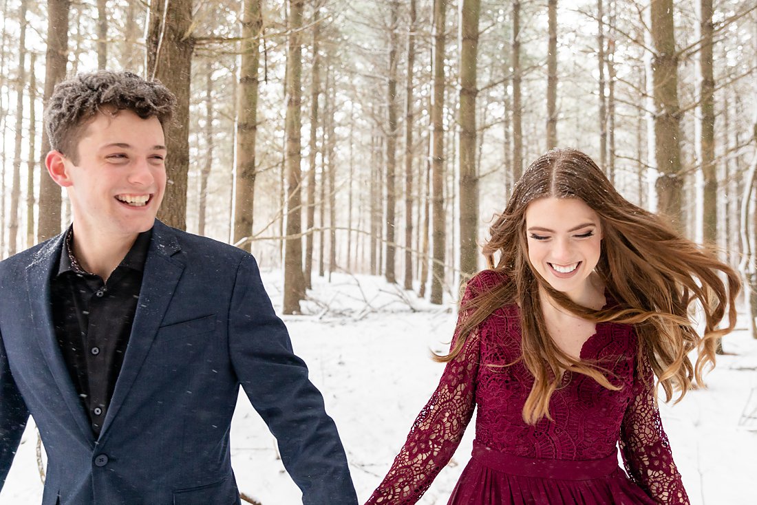 happy couple running in a pine tree forest for their winter engagement session
